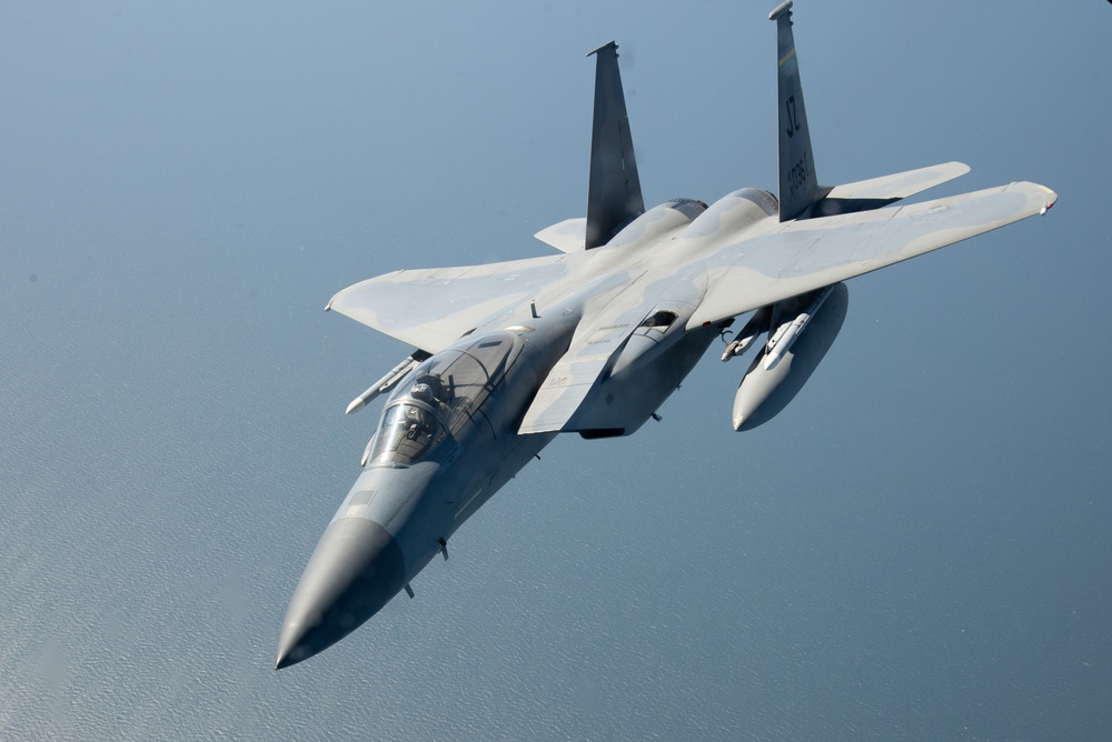 F-15E Strike Eagle in flight, viewed from above showing its full profile against open water, in air superiority gray paint with conformal fuel tanks